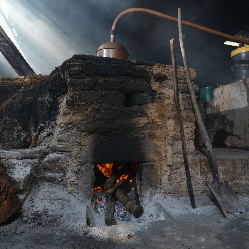 A rustic, stone oven with a roaring fire inside is the focal point, connected to a copper still via a curved pipe. Smoke and steam fill the dimly lit space. A wooden shovel and a long wooden pole lean against the oven. Other distillery equipment, including barrels and containers, are partially visible in the background